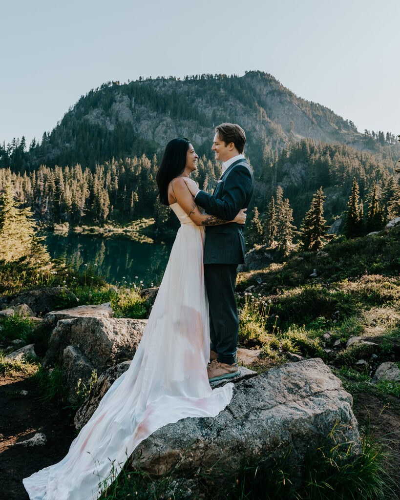 a couple in their wedding attire, embrace while standing on a large rock. In the background, we see a beautiful lake and a mountain, setting the scene for their hiking elopement.