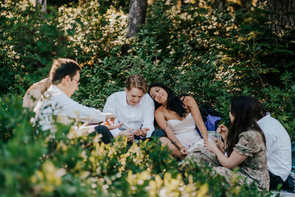 a couple in their wedding attire, sit with their friends in the forest as they share a picnic during their hiking elopement