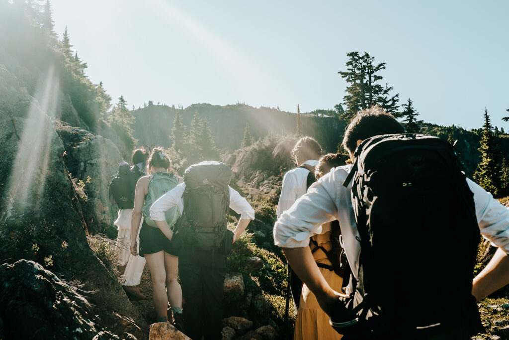 a group of friends with hiking backpacks and wedding attire hike through an exposed ridge line. the sun shines and flares off the camera lens as they explore 