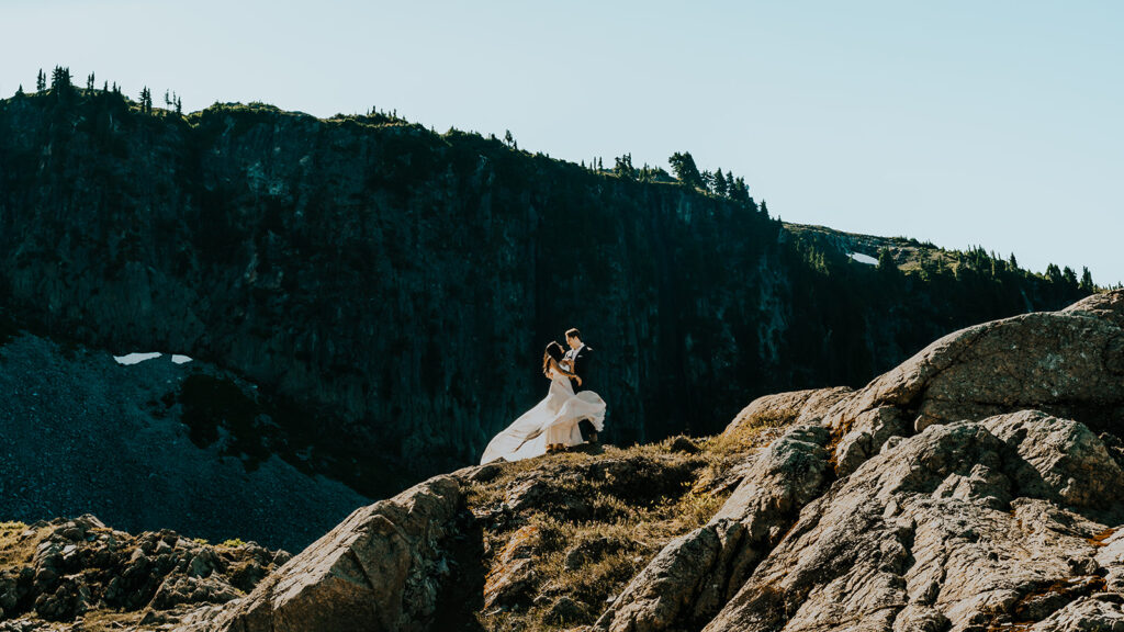 during their hiking elopement, a bride and groom dance on a small cliff with mountains in the background during their hiking elopement