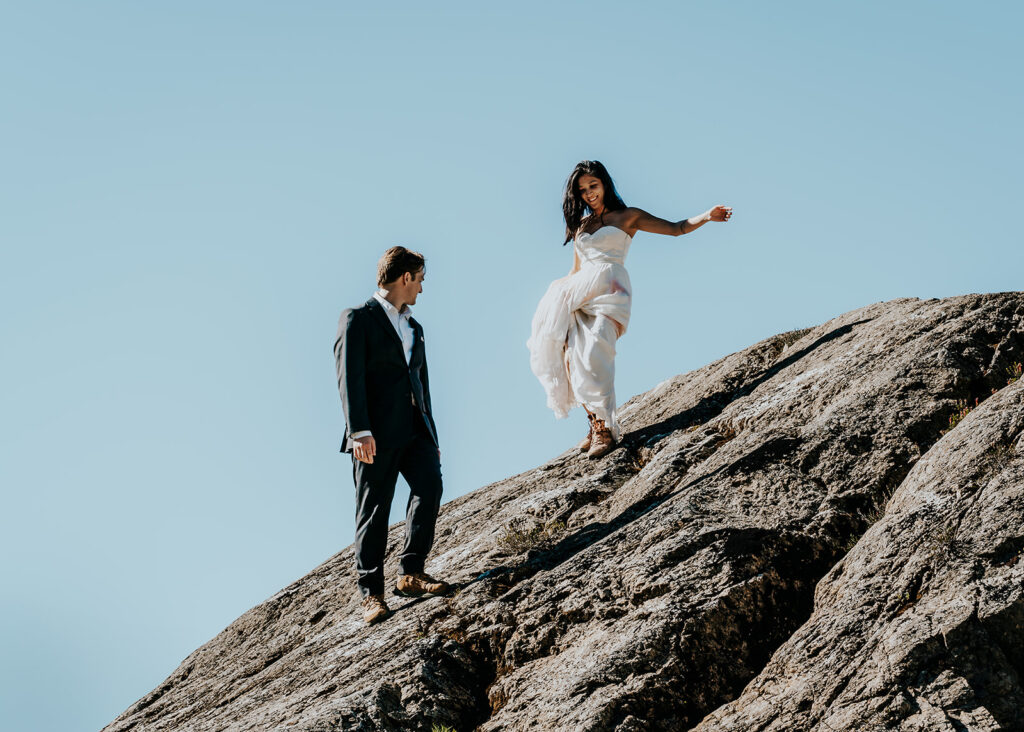 a couple in their wedding attire scrambles down a large boulder during their hiking elopement