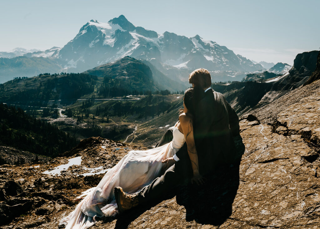 a couple in wedding attire sit on a large, flat rock. they melt into each other as they gaze at a snowy mountain range