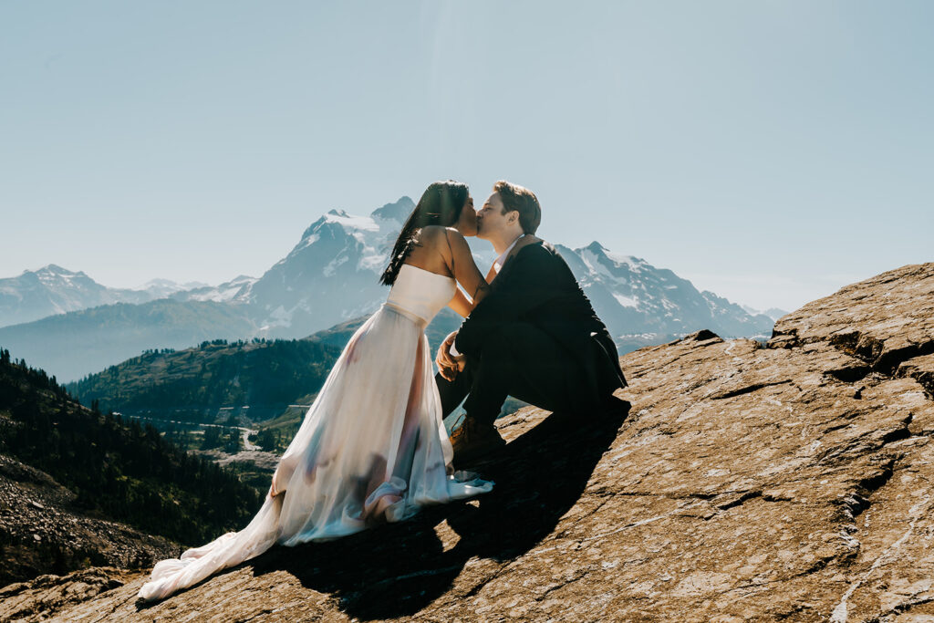 a couple in wedding attire sit on a large, flat rock. they face each other as they kiss. A snowy mountain range serves as their background