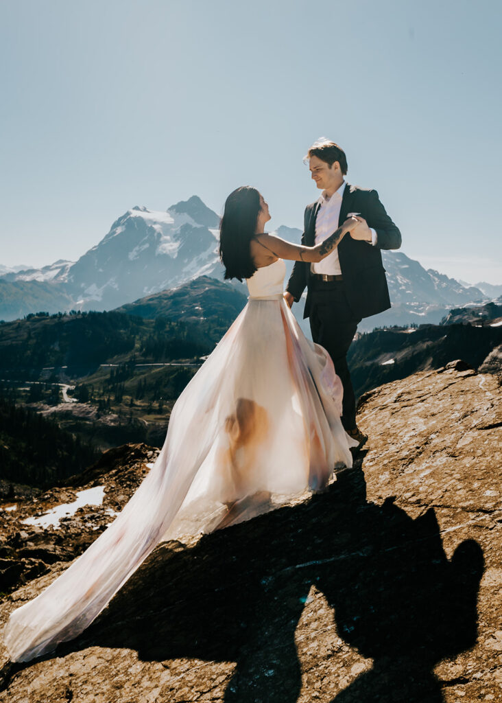 a couple in wedding attire dance on a large, flat rock. they smile at each other with a snowy mountain range in the background