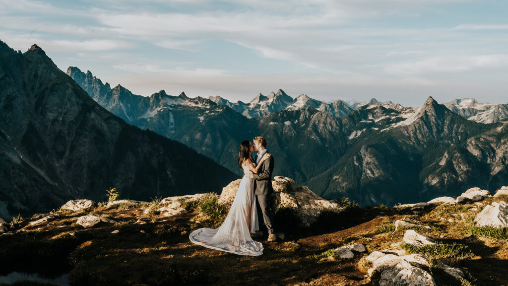 a bride and groom embrace in their wedding attire. A sweeping mountain range serves as their backdrop for this hiking elopement