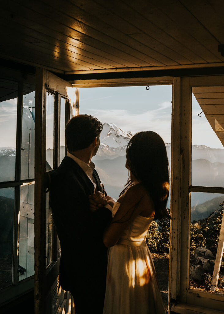 during their hiking elopement, a couple embraces inside a fire tower. they gaze out at a snowy mount bakeer