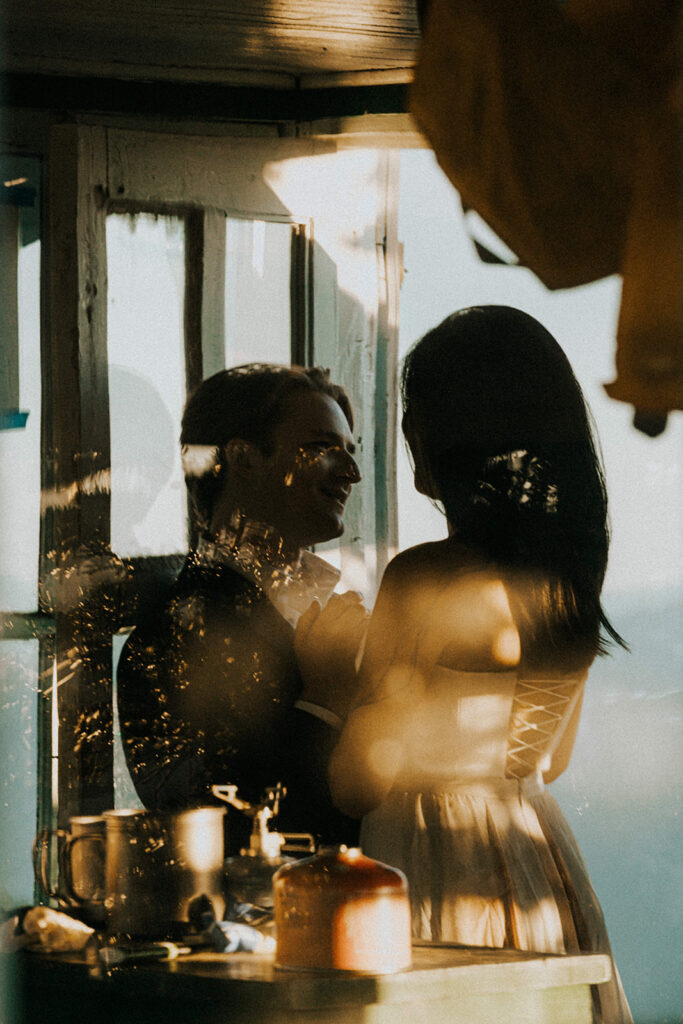 during their hiking elopement, a couple embraces inside a fire tower. The image is taken through the glass adding a romantic and textured foreground 