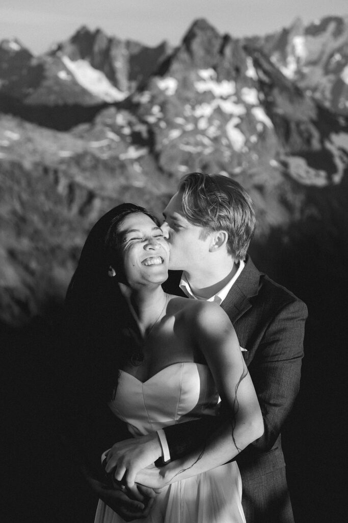 a black and white image of a couple embracing during their hiking elopement. The groom hugs his bride from behind and she smiles widely facing the sun