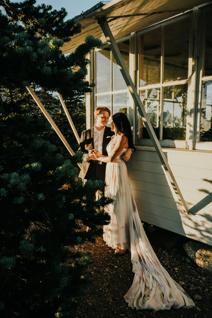 During their hiking elopement, a couple in their wedding attire embrace against the walls of a fire tower. 