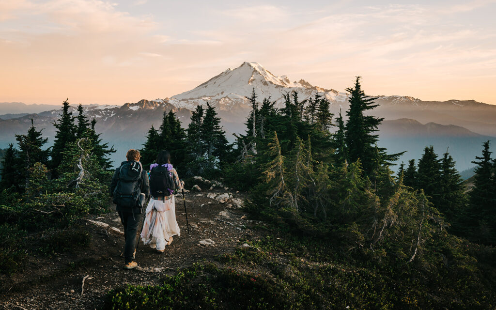 a bride and groom in their wedding attire explore a mountain range during their hiking elopement. mount baker can be clearly seen in the background