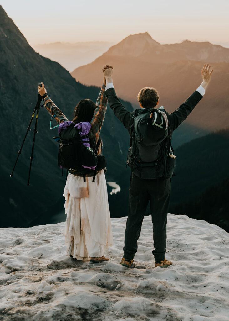 a bride and groom celebrate their hiking elopement while watching a striking sunset. They wear their hiking gear and throw up their hands in excitement