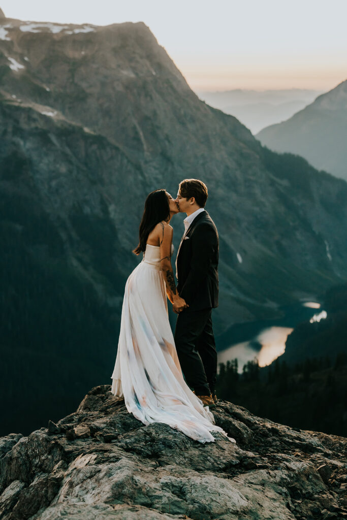 a couple kisses at the top of a mountain. The Cascade range creates a stunning backdrop. They are in their wedding attire during their hiking elopement