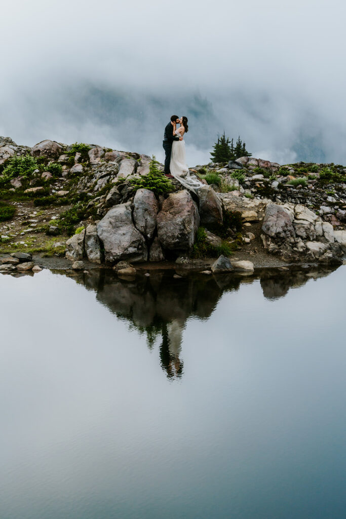 a bride and groom in their wedding attire kiss on the top of a rock, the scene is reflected in the waters of the tarn below them during their North cascades elopement