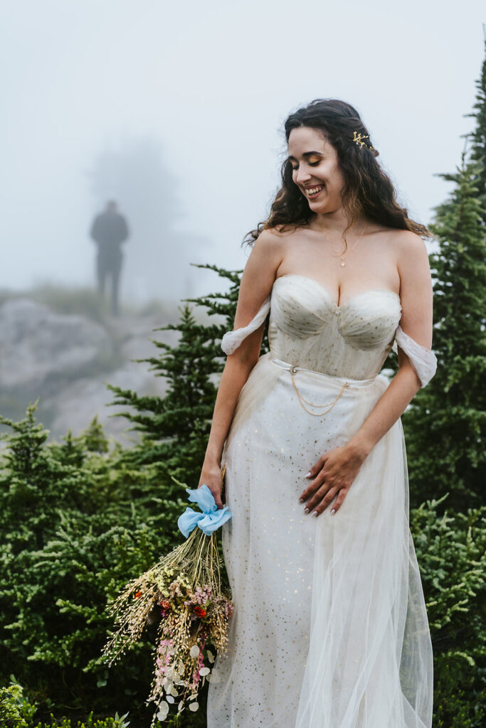 an editorial shot of a bride showing off her wedding dress amongst a foggy landscape. through the fog and in the distance, we see the outline of her groom waiting to see her for the first time during their north cascades elopement