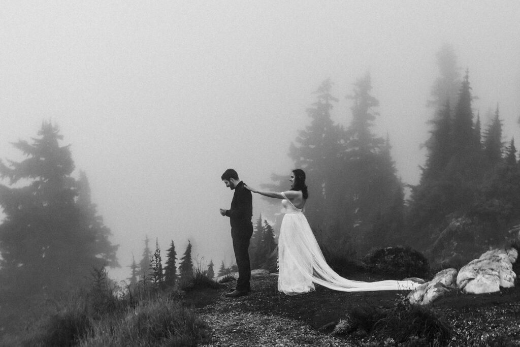 a black and white image of a bride and groom during their first look during their north cascades elopement. this still is just before she turns him around. they are surrounded by beautiful trees and atmospheric fog