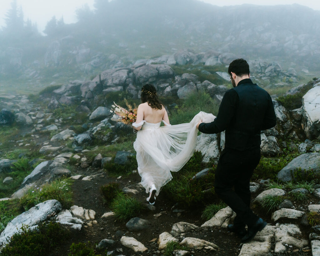 a moody landscape photo. a bride leads the way down a rock trail, her groom carries the train of her wedding dress, while she carries a bright bouquet. Fog surrounds them, but the landscape can still be seen during their North Cascades elopement 