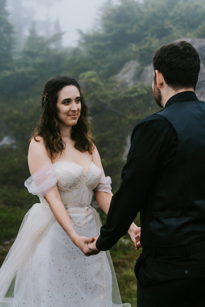 a bride gazes lovingly at her groom during their north cascades elopement ceremony. they are surrounded by fog and greenery, giving an ethereal vibe 