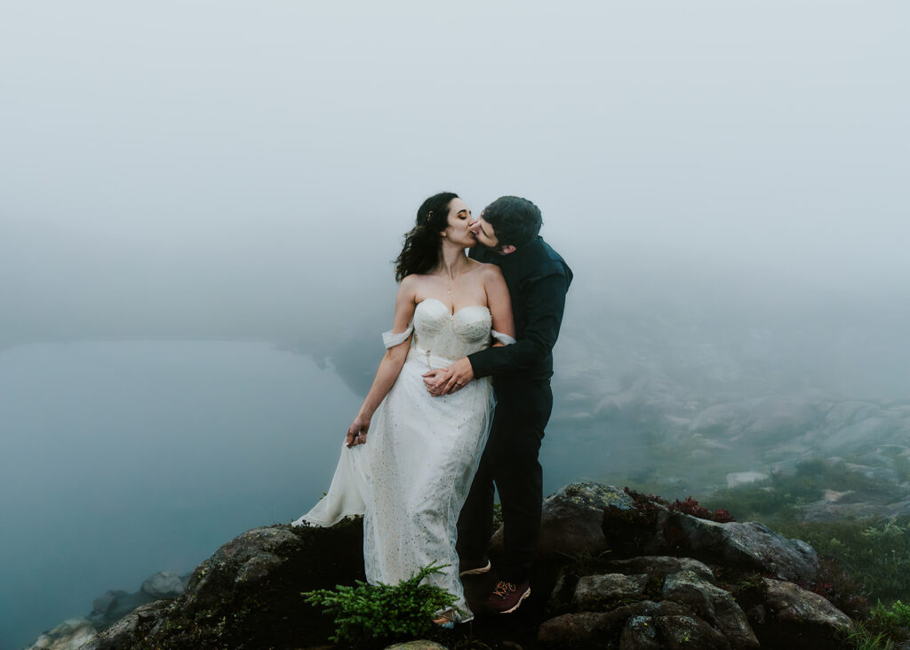 a moody landscape shot with deep blues and green. a couple kisses in a rocky outcropping. The fog swirls around them and we can barely see the small tarn below them. 