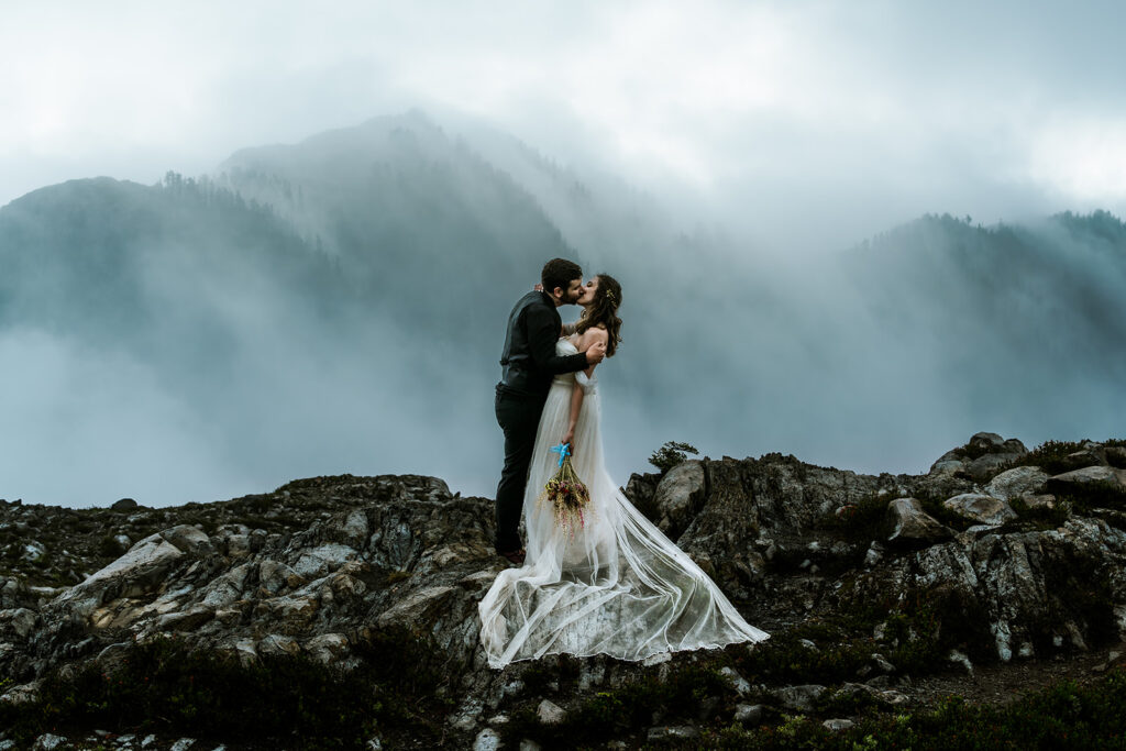 A moody, dramatic landscape image of a bride and groom kissing as they stand on a rocky outcropping. The soft image of mountains can be seen through the misty fog that surrounds them during their North Cascades elopement. 