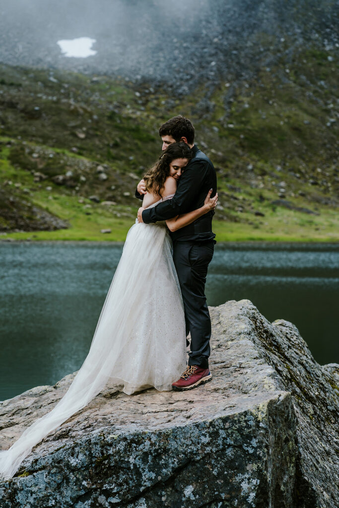 a couple in wedding attire stand on a large granite slab and embrace deeply. an alpine lake can be seen shimmering behind them and a foggy mountain create a mystical view 