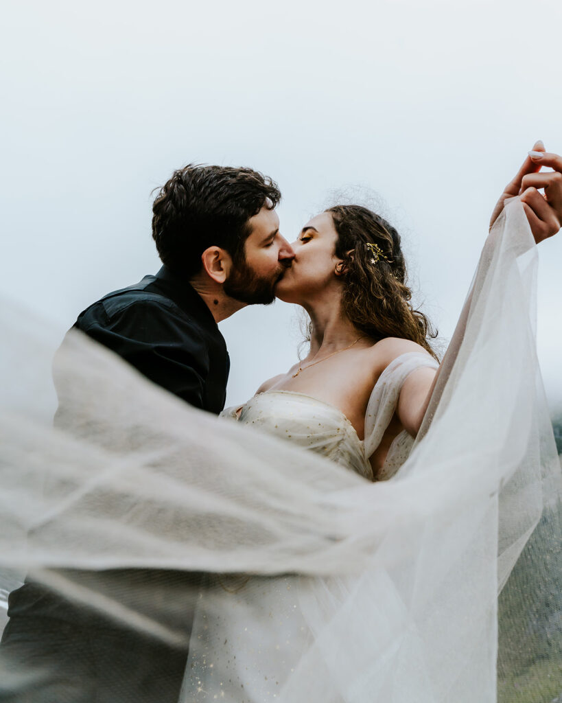 shot from below, a bride and groom kiss framed by a foggy sky and the swirling fabric of her dress during their north cascades elopement