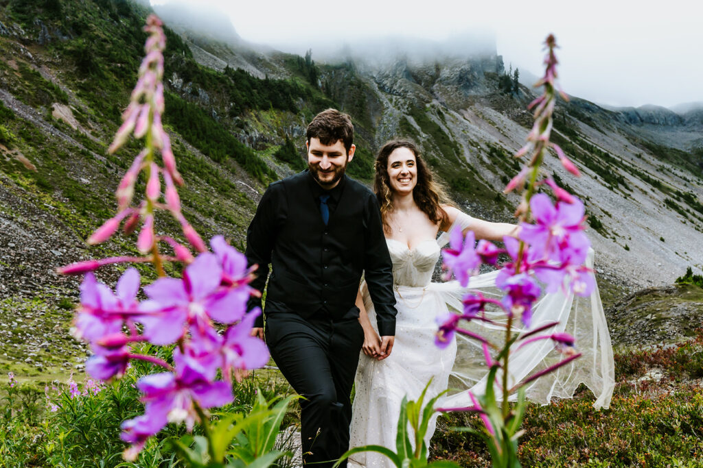 A bride and groom in their wedding attire smile widely as they walk through a green landscape. Mountains rise up behind them and purple, alaskan wildfire frames them as they walk towards the camera. The bride spreads the fabric of her dress, feeling the wind that blows past them.