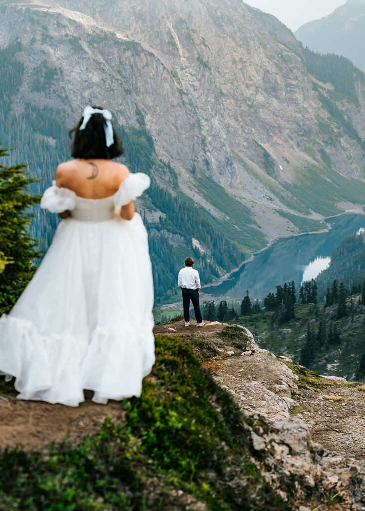 a before moment of a couples first look during their north cascades elopement. he stands waiting on the edge of a mountain as she walks towards him 