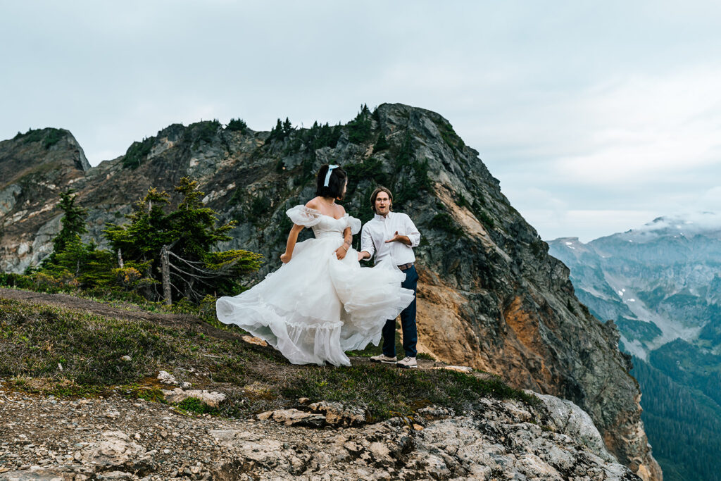 during their north cascades elopement a bride shows off her wedding dress to her groom. he smiles widely as she twirls with the mountains in the frame them in the background