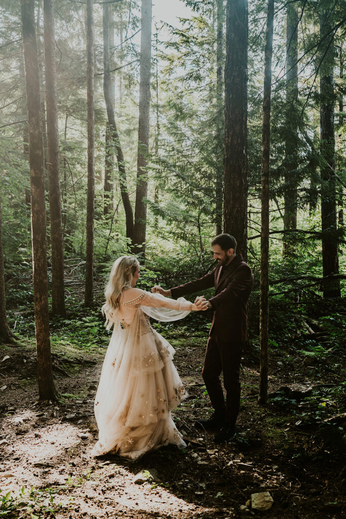 in the forest, a bride and groom hold hands and spin. golden light filters through the green canopy making them glow