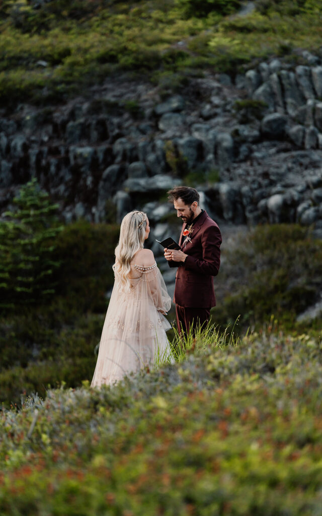 during their north cascades elopement, a bgroom reads his vows to his bride. wildflowers paint the foreground and granite walls of the mountains create the backdrop for this moment