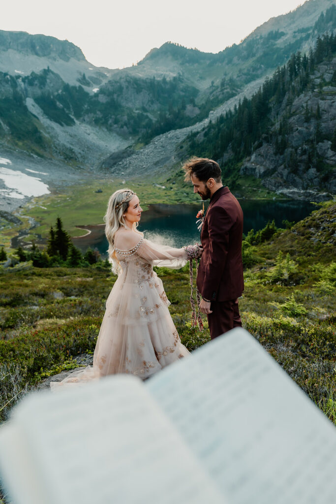 a perspective shot from the officiants pov of a couple during their north cascades elopement ceremony. they are doing a hand fasting ceremony with an alpine lake and stunning mountains in the background 