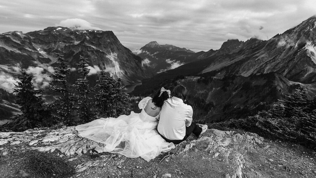 a bride and groom sit on the edge of a cliff and gaze out at the rolling mountains during their north cascades elopement