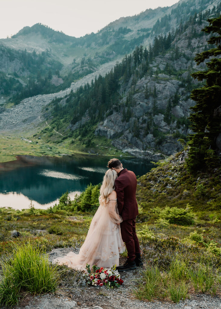 During their North Cascades elopement, a couple in wedding attire embrace and gaze out at a blue lake surrounded by rolling mountains and deep, green trees. they rest thier heads on each other, sweetly as they take in the view. 