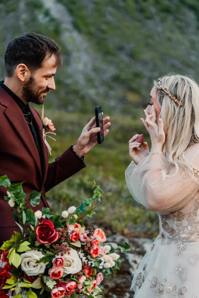 a groom holds a phone for his bride to use as a mirror as she fixes her makeup after crying during their north cascades elopement ceremony 