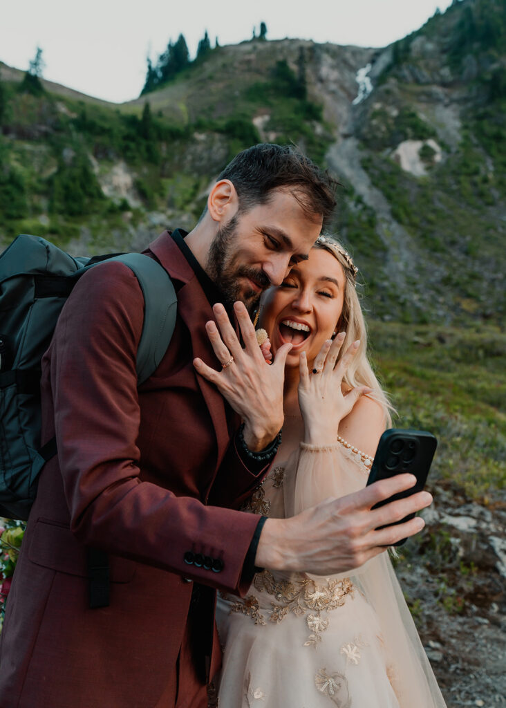 a couple in their wedding attire smile widely, holding up their hands to showcase their wedding rings as they take a selfie to celebrate their north cascades elopement