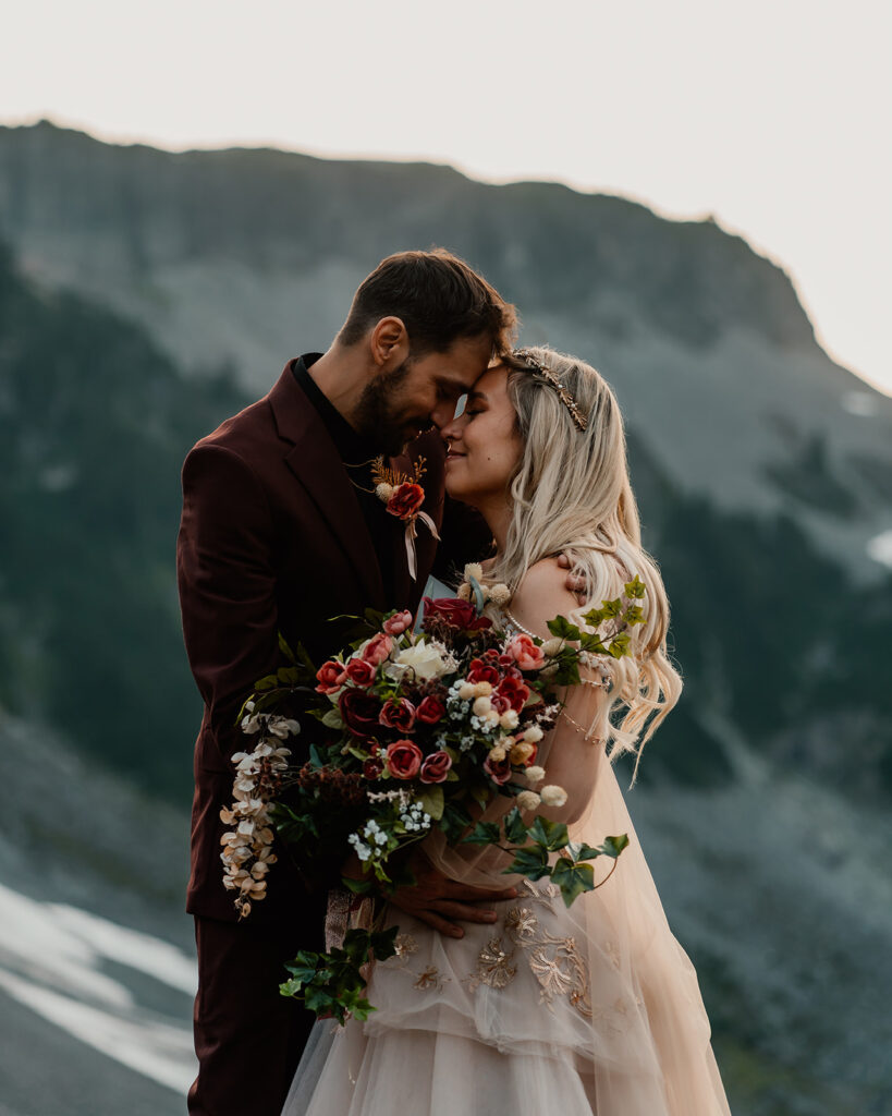 A couple in wedding attire embrace and snuggle their faces together during their North Cascades elopement. The bride holds a cascading red and pink bouquet. The groom holds her. Blue mountains and tiny patches of snow can be seen behind them.