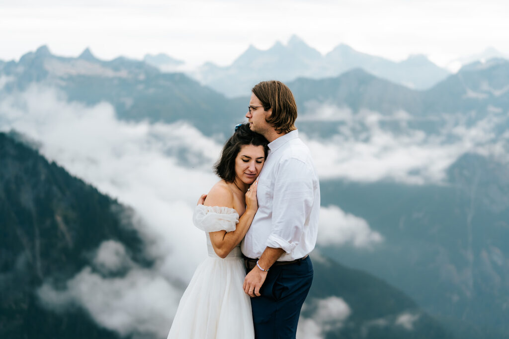 a bride rests her head and hands on her grooms chest. Blue mountains, with rolling clouds create a magical backdrop for their North Cascades elopement 