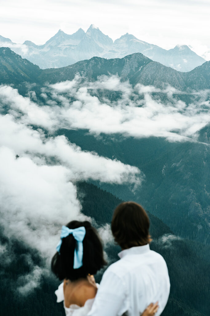 a bride and groom gaze out at the blue mountains before them as white clouds begin to roll in through the peaks. the back of their heads are blurred framing the scene of their north cascades elopement