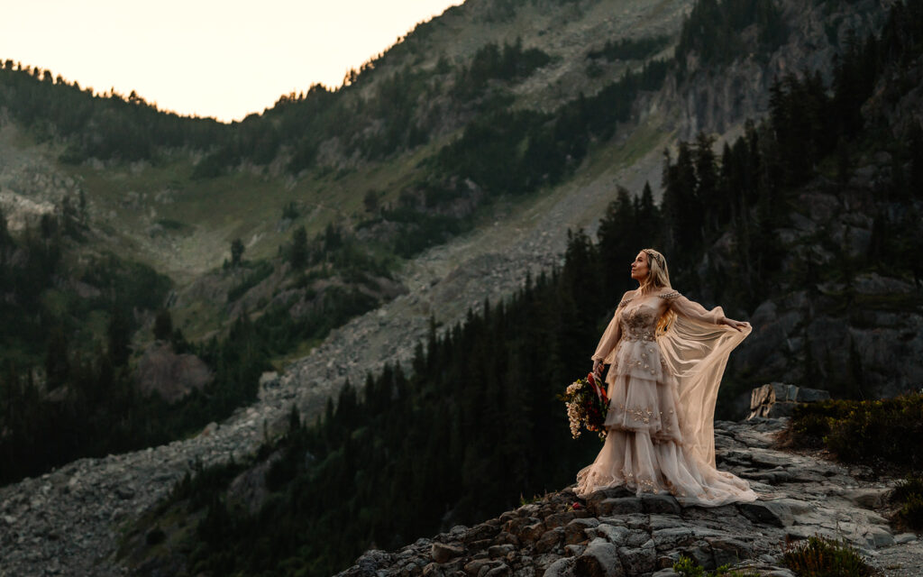 An ethereal landscape portrait of a bride in her wedding dress. she flows the fabric of her dress as she gazes out at the surrounding mountains. Her bouquet hangs in her hands. the light is soft and golden during her north cascades elopment  