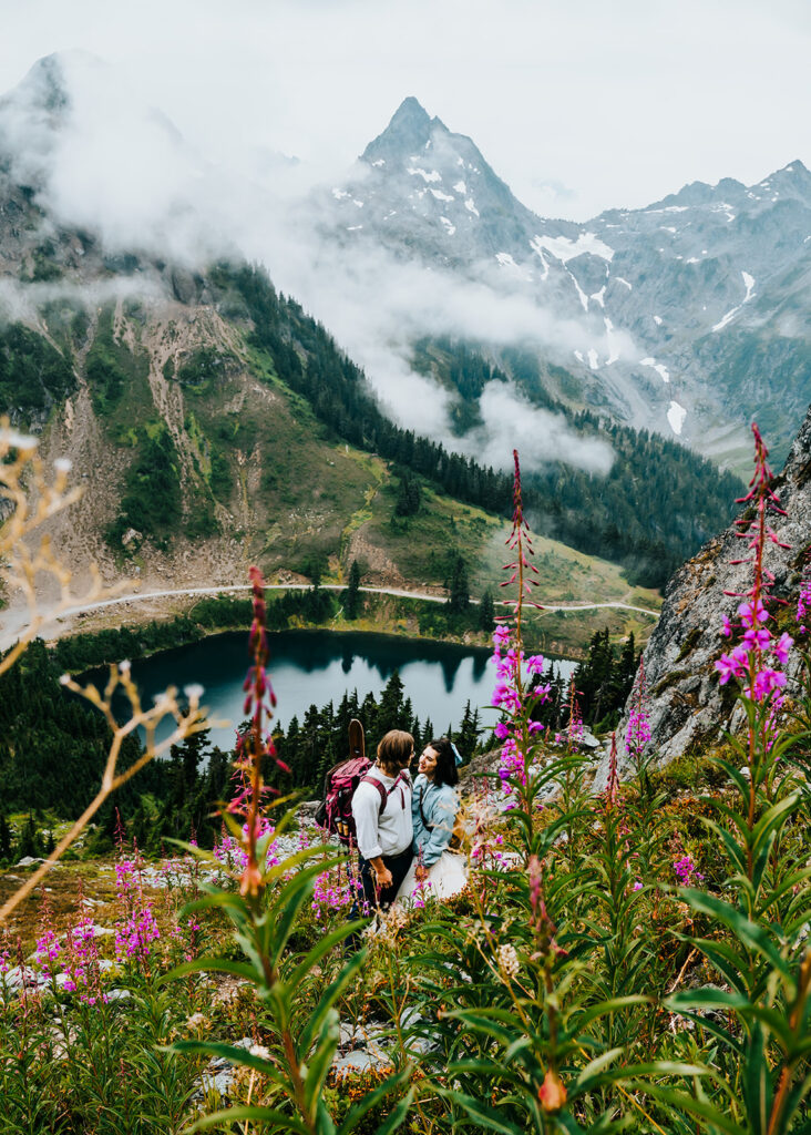 a couple in their wedding attire hike down a wildflower filled trail. the gaze at each other with the mountains rising behind them and a glassy lake below them. They are framed by bright purple alaskan fireweed during their North Cascades elopement