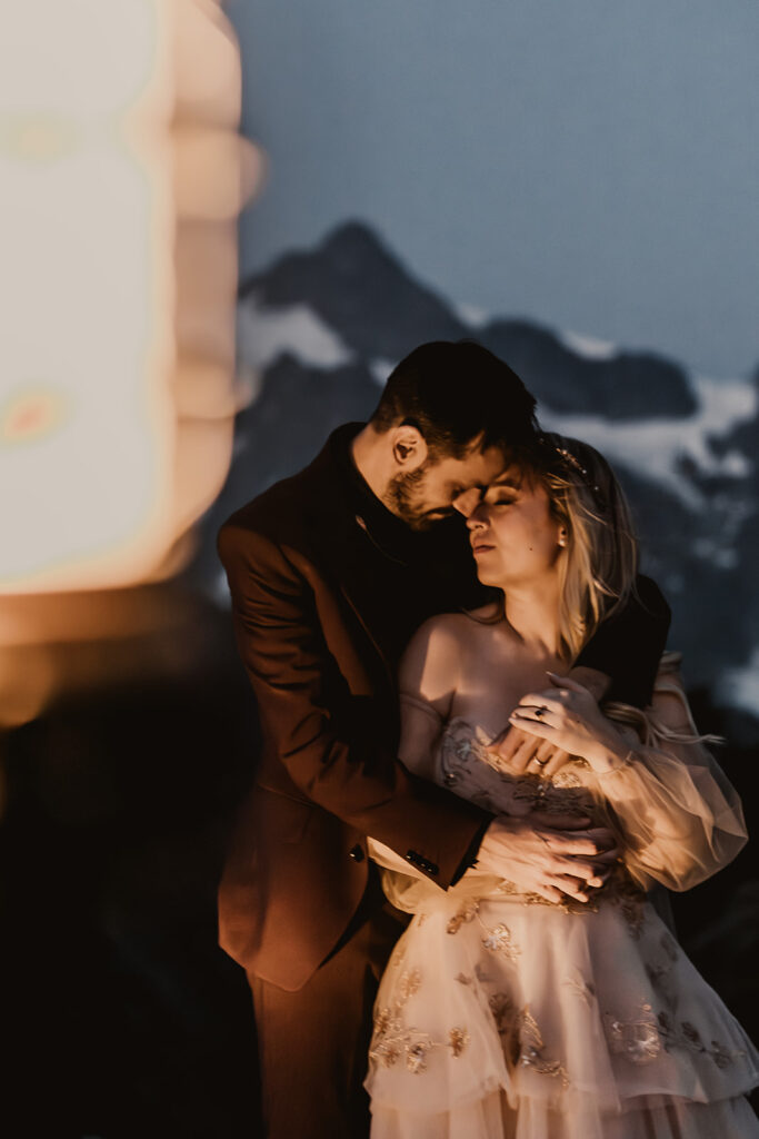 during duck, a couple in their wedding attire embrace romantically. their faces meet, and their hands intertwine around the brides body. snowy, jagged mountain peaks create a their background and the soft glow of a lantern can be seen in the foreground. 