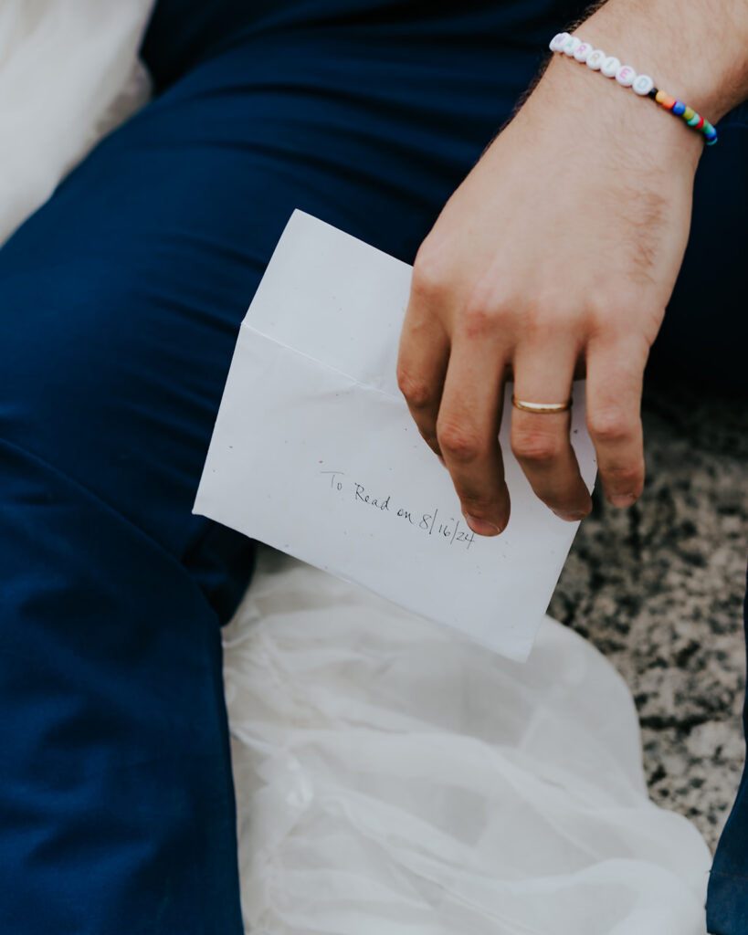 a detail shot of a groom with his new wedding band on his ring finger. he holds an envelope that reads "to read on 8/16/24" their north cascades elopement date