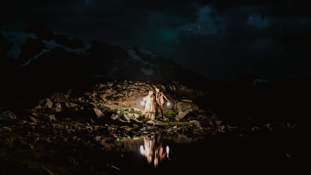 a magical landscape shit of a starry night sky, a faint outline of a snowy mountain and a couple kissing as they hold glowing lanterns during their north cascades elopement