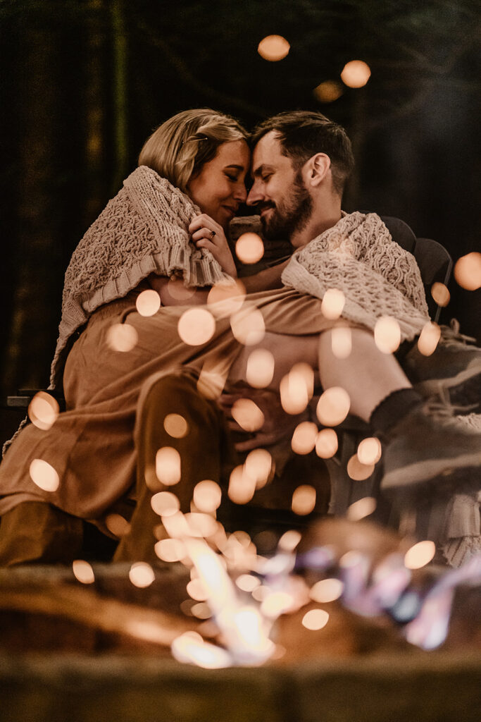 after their North Cascades elopement, a couple sit in front of a glowing campfire. The bride sits in her husbands lap and embers sparkle in the foreground. They press their faces together ad smile softly as they wrap up in a beige, knitted blanket. 