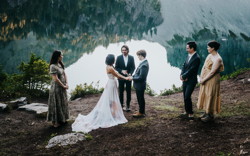 a bride and groom stand on the shores of an alpine lake surrounded by guests as they exchange vows 