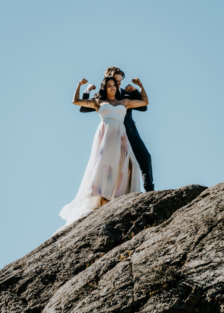 a bride and groom in wedding attire goof around during their north cascades elopement and flex their arms while standing on top of a boulder they just scaled