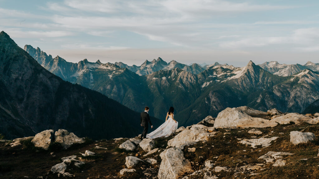 A couple in wedding attire walk a long a path of a rock outcropping. Blue, jagged peaks jut out behind them during their North Cascades elopement.