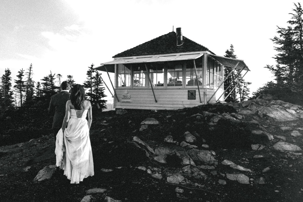 a black and white image of a couple in their wedding attire walking towards a fire lookout during their north cascades elopement