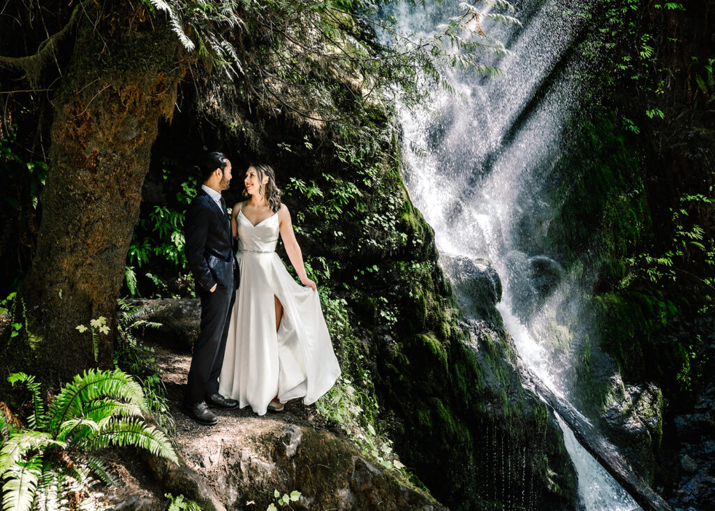 a groom and bride gaze lovingly at each other in front of a waterfall. the water from the falls behind them glitter in the sunlight. They are dressed in their wedding attire to prepare for their ruby beach elopement