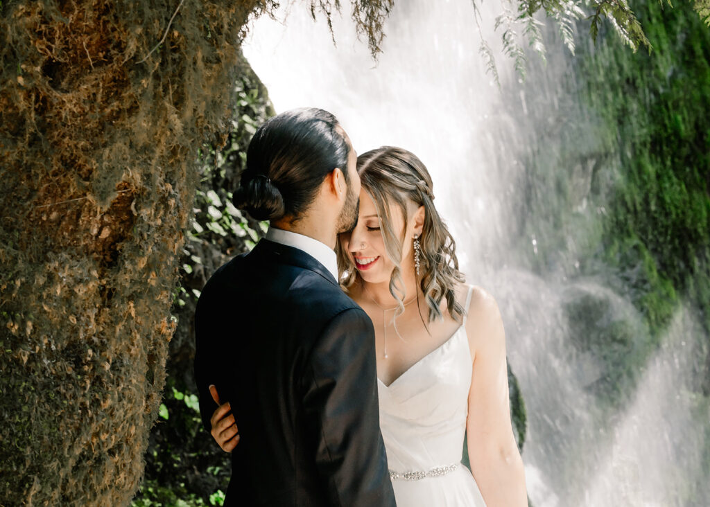 a groom kisses his brides forehead as water from the falls behind them glitter in the sunlight. They are dressed in their wedding attire to prepare for their ruby beach elopement